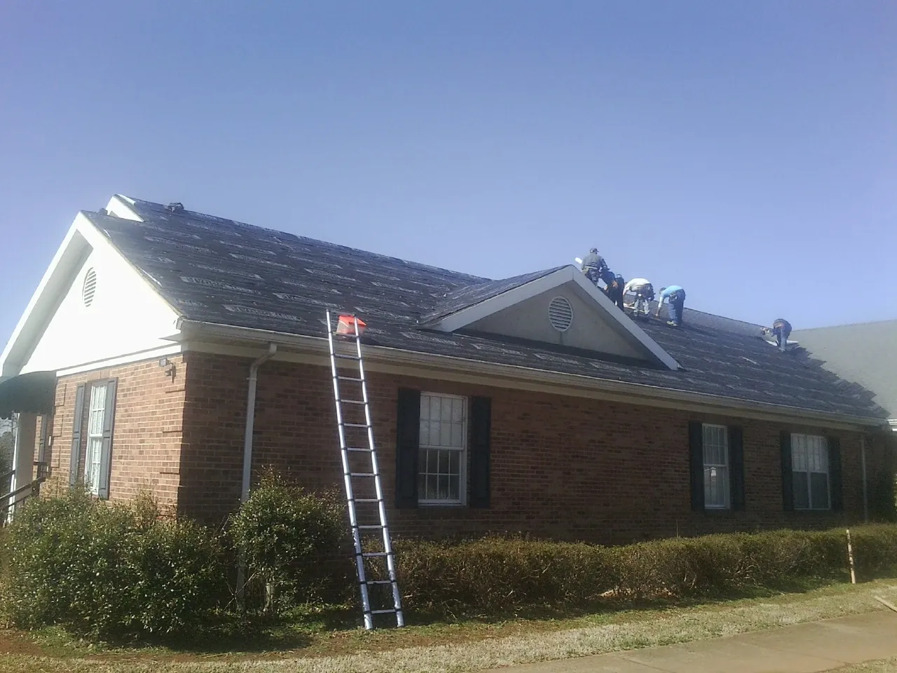 Crew tearing shingles off a brick ranch roof with a ladder set — Metal Roof Repair work by our crew in Montross