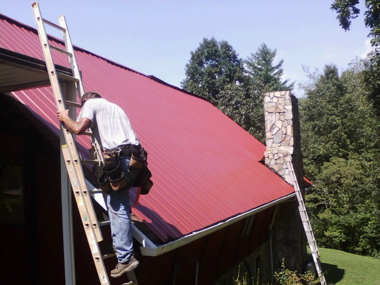 Roofer on a ladder beside a red metal roof and stone chimney — Soffit & Fascia Repair by Roofing Brothers in Montross